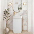 A modern bathroom vanity setup featuring a white ribbed pedestal sink with a gold faucet, set against a white marble archway wall with an oval mirror and two round wall lights. A small potted eucalyptus plant in a ceramic vase sits to the left.