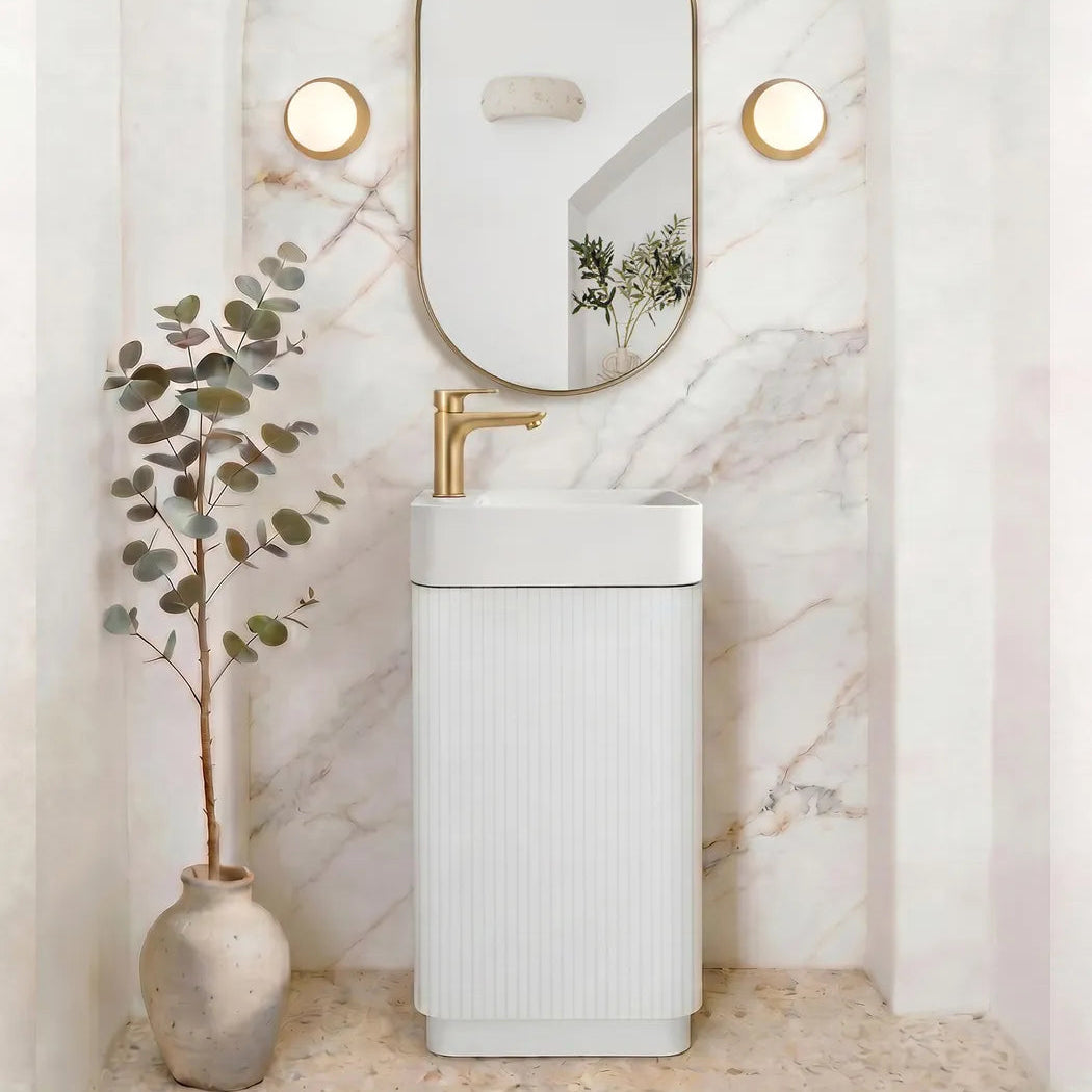 A modern bathroom vanity setup featuring a white ribbed pedestal sink with a gold faucet, set against a white marble archway wall with an oval mirror and two round wall lights. A small potted eucalyptus plant in a ceramic vase sits to the left.