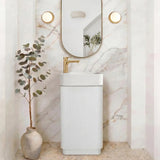 A modern bathroom vanity setup featuring a white ribbed pedestal sink with a gold faucet, set against a white marble archway wall with an oval mirror and two round wall lights. A small potted eucalyptus plant in a ceramic vase sits to the left.