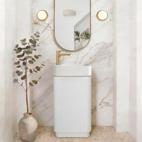 A modern bathroom vanity setup featuring a white ribbed pedestal sink with a gold faucet, set against a white marble archway wall with an oval mirror and two round wall lights. A small potted eucalyptus plant in a ceramic vase sits to the left.
