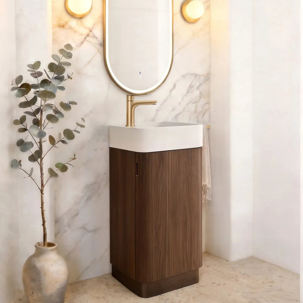 A modern bathroom vanity featuring a tall dark wood cabinet, a white rectangular basin, a gold faucet, and an oval gold-framed mirror mounted on a white marble arched wall. A potted eucalyptus branch stands on the floor to the left.