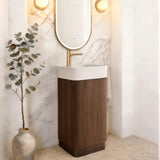 A modern bathroom vanity featuring a tall dark wood cabinet, a white rectangular basin, a gold faucet, and an oval gold-framed mirror mounted on a white marble arched wall. A potted eucalyptus branch stands on the floor to the left.