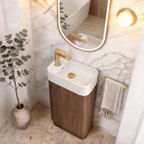 A modern bathroom vanity setup featuring a rectangular white sink with a gold faucet, mounted on a dark wood cabinet. Above it is an oval mirror with a gold frame, flanked by two warm wall lights.