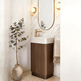 A modern bathroom vanity featuring a white ceramic sink, a dark brown wood cabinet, a gold faucet, and an oval mirror with a gold frame, set against a marble-look wall with warm wall sconces.