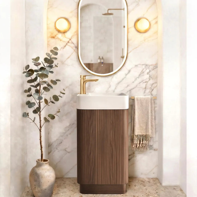 A modern bathroom vanity setup featuring a rectangular white sink with a gold faucet, mounted on a dark wood pedestal. Above it is an oval mirror with a gold frame, set against a marble-patterned wall with built-in round lights.