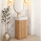 A modern bathroom vanity featuring a light oak wood cabinet, a white rectangular sink, a gold faucet, and an oval mirror with a gold frame, set against a white marble archway wall with warm wall lights.