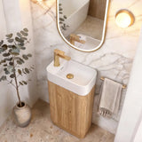 A modern bathroom vanity featuring a white ceramic sink basin with a light wood cabinet base, a gold faucet, and an oval mirror with a gold frame. The setup is styled in a warm, minimalist bathroom with marble walls, a gold wall mirror light, and a potted eucalyptus plant.