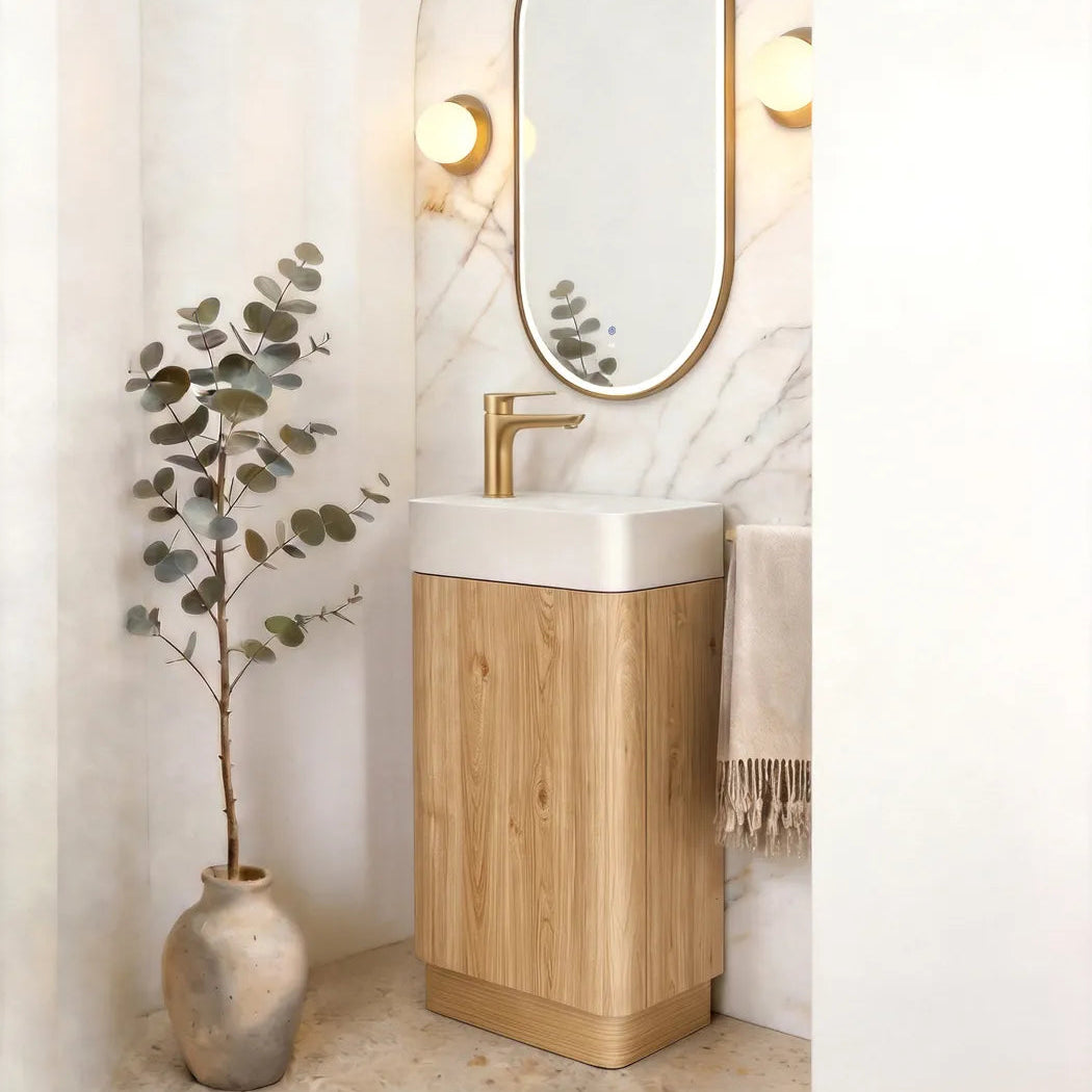 A modern bathroom vanity featuring a light oak wood cabinet base, a white ceramic sink, a gold faucet, and an arched wall mirror with a gold frame. The setup is styled in a warm, minimalist bathroom with a marble accent wall, a potted eucalyptus plant, and a beige towel.