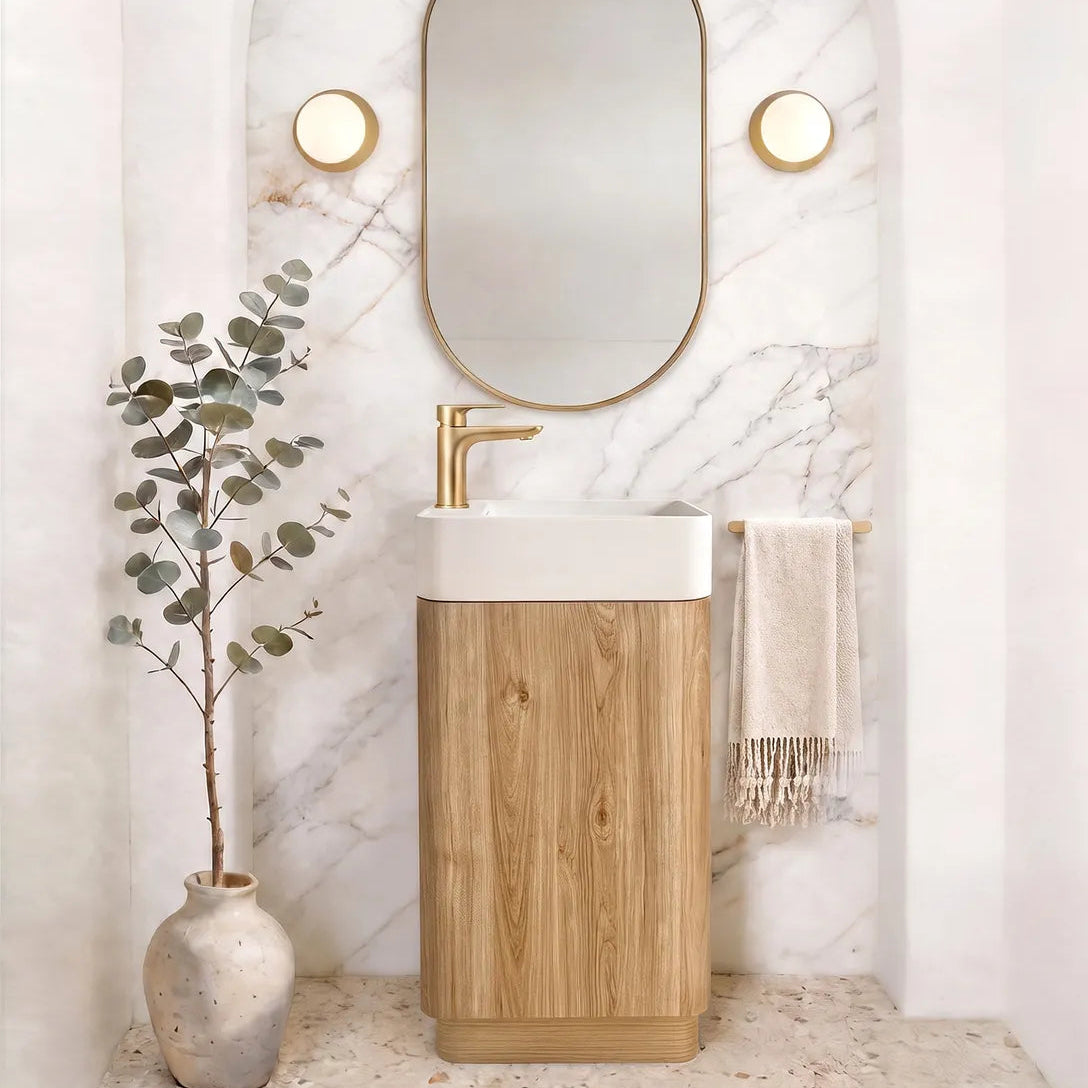 A modern bathroom vanity setup featuring a rectangular white sink basin mounted on a light oak wood cabinet, with a gold faucet. Above it is an arched white marble wall with a gold-framed oval mirror and two round gold wall sconces.