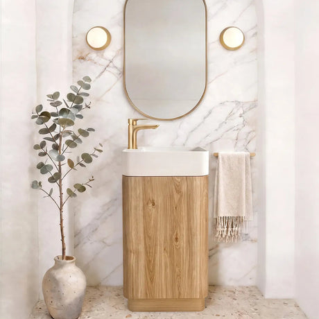 A modern bathroom vanity setup featuring a rectangular white sink basin mounted on a light oak wood cabinet, with a gold faucet. Above it is an arched white marble wall with a gold-framed oval mirror and two round gold wall sconces.