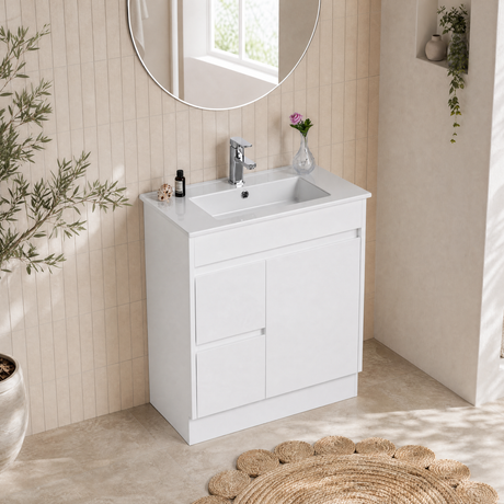 A white bathroom vanity cabinet with an integrated white sink and a chrome faucet, shown in a bright, modern bathroom setting with a round mirror, tiled wall, and woven rug.
