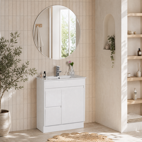 A modern white bathroom vanity cabinet with an integrated white sink and chrome faucet, set against beige tiled walls in a bright, sunlit bathroom. A round wall mirror hangs above, and a potted plant sits to the left.