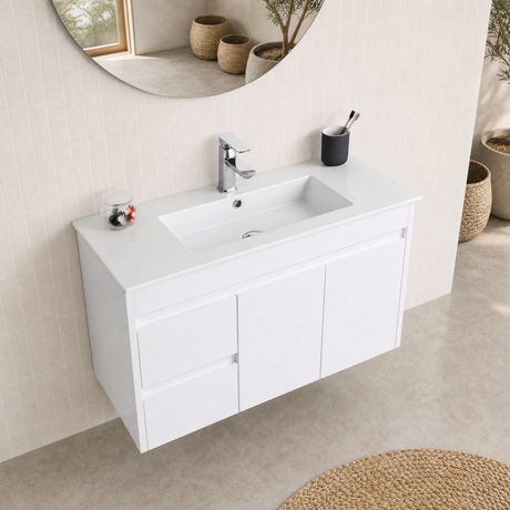 A modern white wall-mounted bathroom vanity with an integrated rectangular sink, chrome faucet, and three cabinet doors, shown in a clean beige-toned bathroom setting with a round mirror and small decor items.