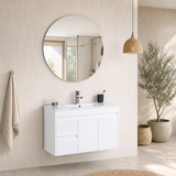 A modern bathroom vanity setup featuring a white wall-mounted cabinet with an integrated white sink, a round backlit mirror, a chrome faucet, and a black cup holder, set against a beige tiled wall with a hanging towel and a potted plant.