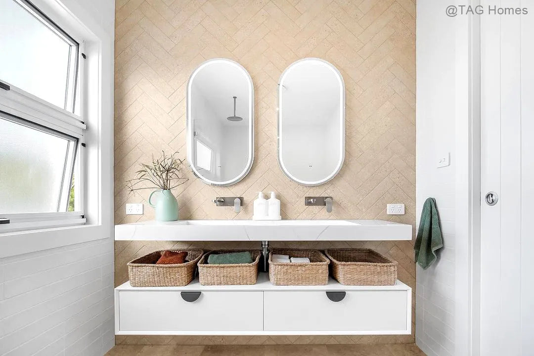Bathroom vanity with two mirrors, baskets, and toiletries against a beige wall.