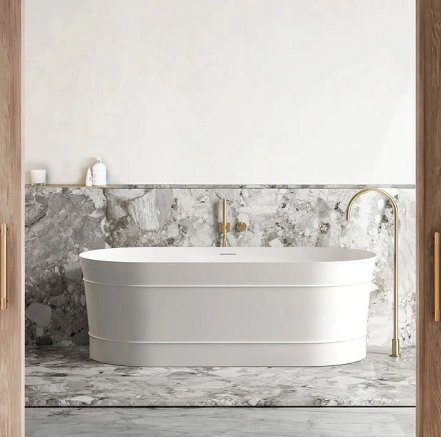 White freestanding bathtub in a bathroom with marble walls and gold fixtures.