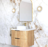 Bathroom vanity with wooden cabinet, white sink, and gold faucet against a marble wall.