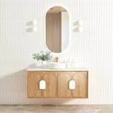 Bathroom vanity with wooden cabinet, round mirror, and decorative plant against a light tiled wall.