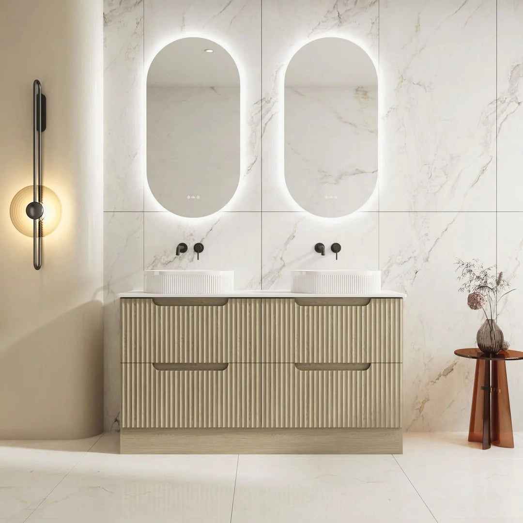Bathroom vanity with double sink and illuminated mirrors against a marble wall.