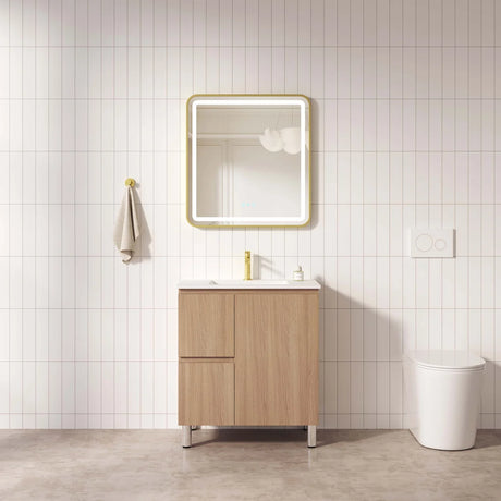 A modern bathroom vanity set featuring a light wood cabinet with a white sink basin, a gold faucet, a gold-framed illuminated mirror, and a white toilet, set against a white tiled wall.