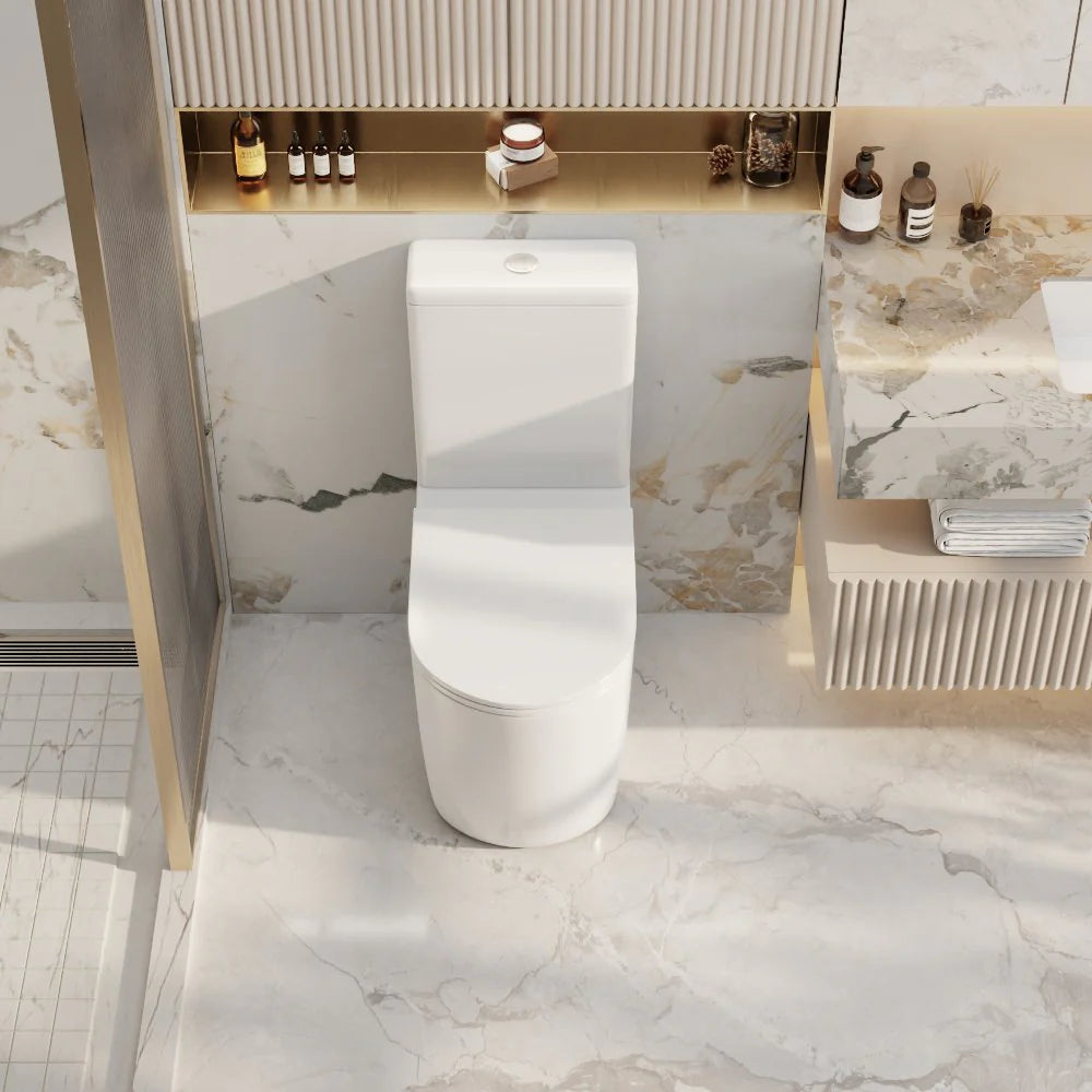 A modern white ceramic one-piece toilet centered in a luxurious bathroom setting with marble walls and floor, a gold-framed shower door, and a matching marble vanity.